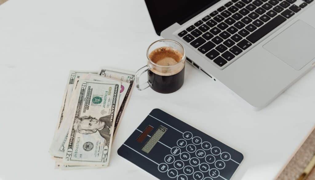 Calculator, notebook, banknotes and a cup of coffee arranged on a desk beside a laptop while budgeting for training tuition