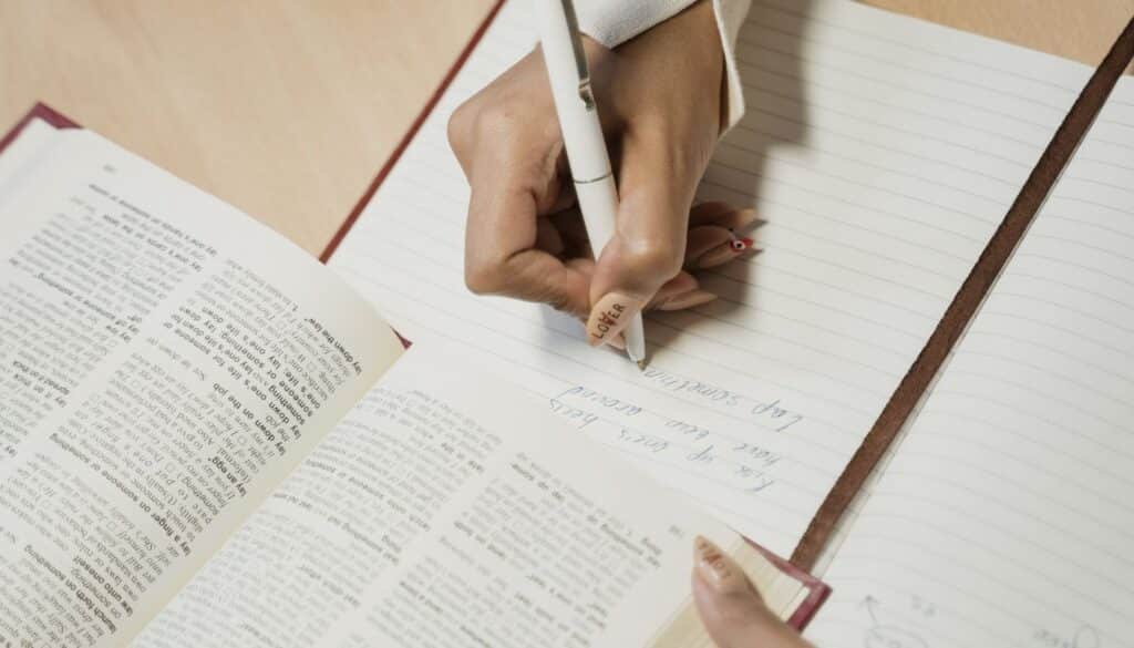 Close up of a person writing notes in a spiral notebook beside open reference books while studying Ayurveda coursework