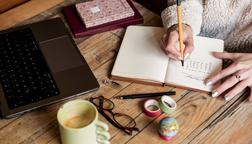 Woman writing figures in a notebook next to a laptop and a mug of coffee while weighing up an Ayurveda training decision